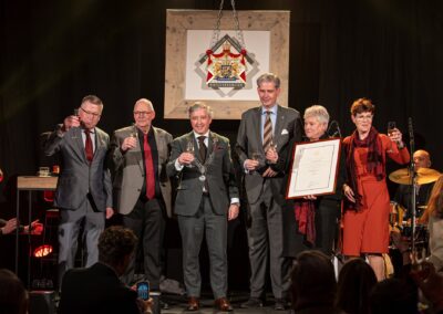 Zes personen heffen het glas op het podium tijdens de uitreiking van het predicaat Hofleverancier aan Verwaal Transport, met de burgemeester in ambtsketen en het wapenschild op de achtergrond