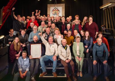 Grote groepsfoto van het team en genodigden op het podium tijdens het Hofleverancier-feest, met de oorkonde en het wapenschild