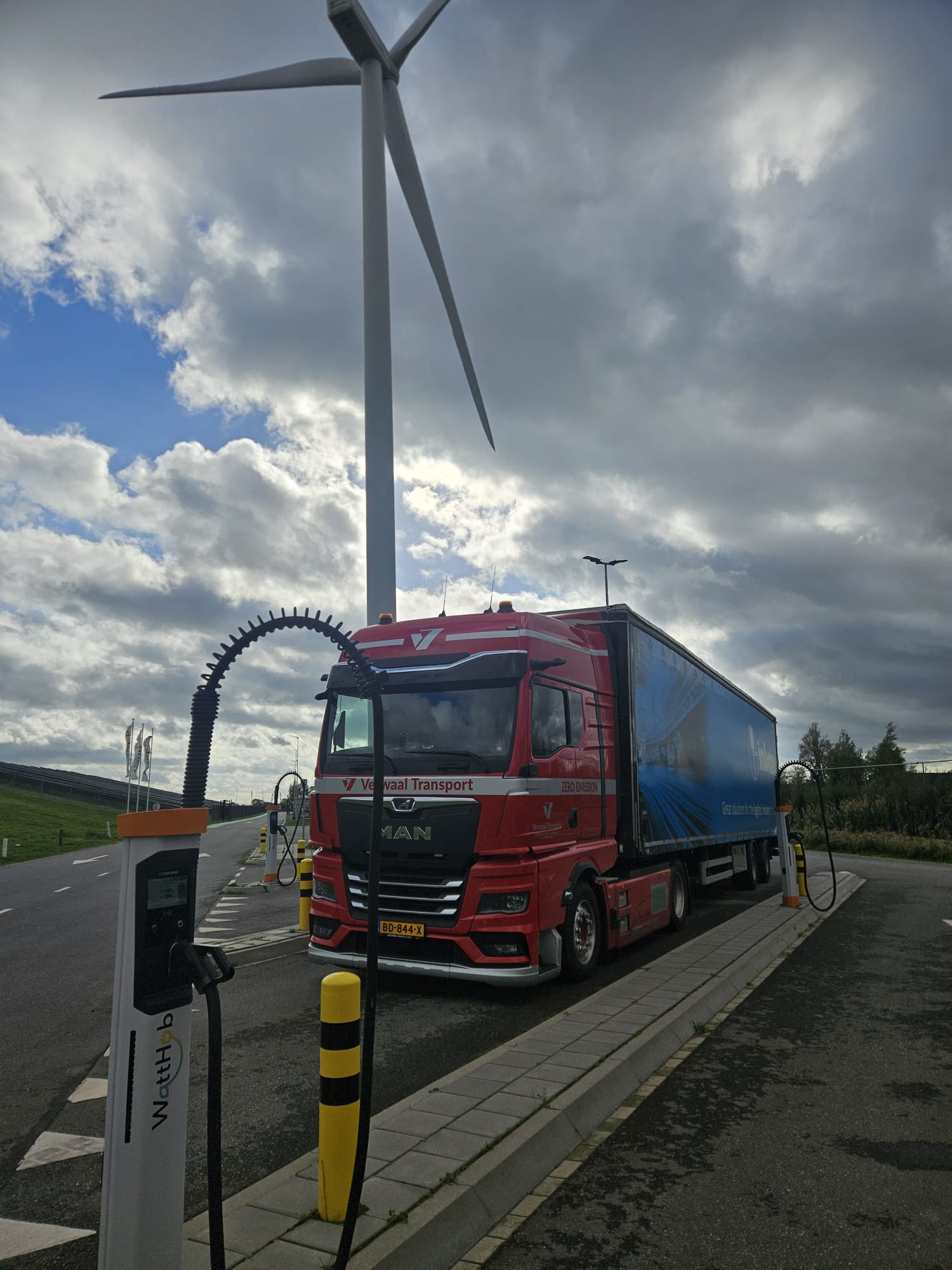 Elektrische MAN vrachtwagen van Verwaal Transport laadt op bij een snellaadpunt met windmolen op de achtergrond